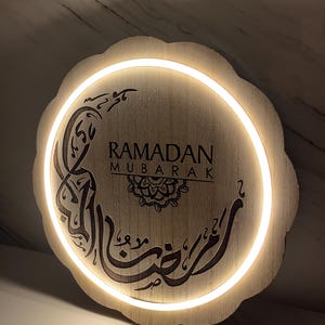Op de afbeelding: Ronde houten Ramadan Mubarak-bord met een geschulpte rand. Het bord toont de woorden "RAMADAN MUBARAK" en Arabische kalligrafie, verlicht door een warmwitte LED-verlichting. Het bord is gemaakt van lichtgekleurd hout.