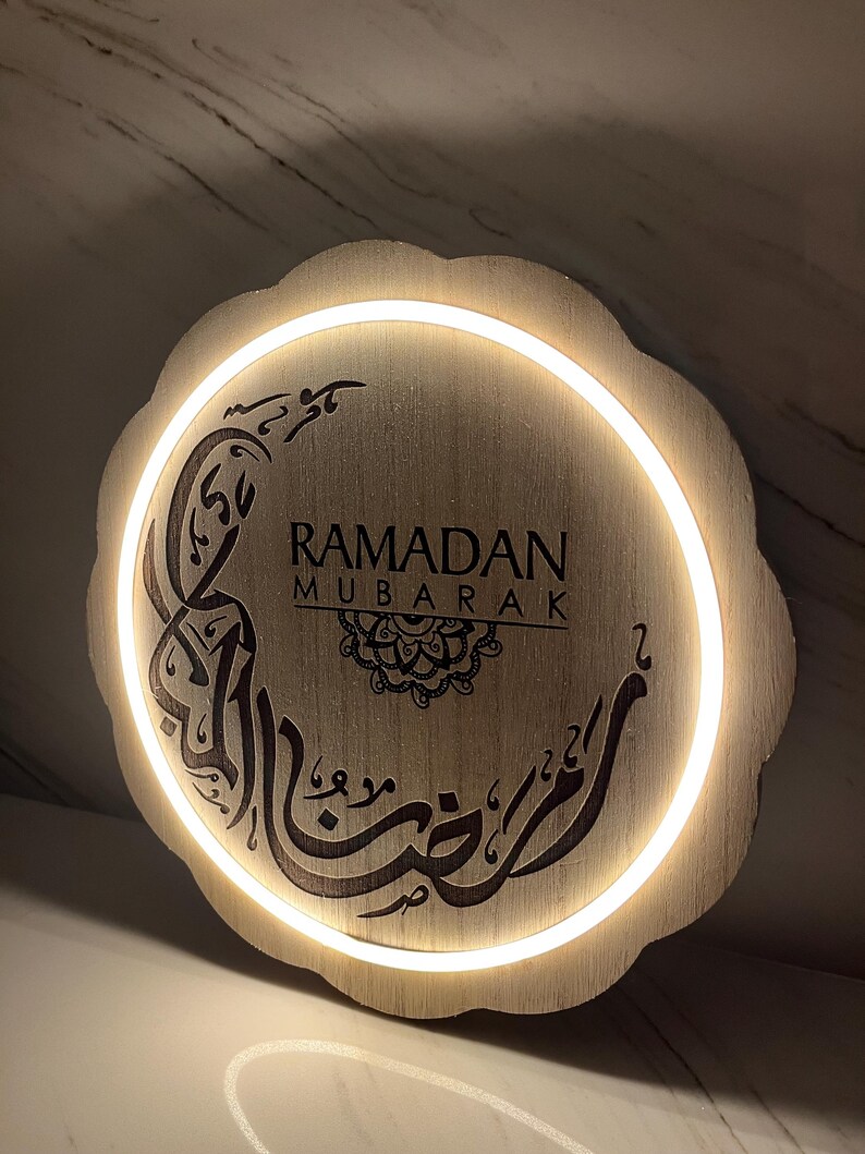 Op de afbeelding: Ronde houten Ramadan Mubarak-bord met een geschulpte rand. Het bord toont de woorden "RAMADAN MUBARAK" en Arabische kalligrafie, verlicht door een warmwitte LED-verlichting. Het bord is gemaakt van lichtgekleurd hout.