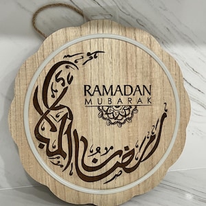 Op de afbeelding: Houten Ramadan Mubarak-bord met een geschulpte rand. Het bord toont Arabische kalligrafie en de woorden "Ramadan Mubarak" in een decoratief lettertype. Een wit cirkelvormig licht omlijst het ontwerp en een jutetouw is bevestigd om op te hangen.