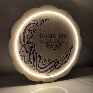 Op de afbeelding: Ronde houten Ramadan Mubarak-bord met een geschulpte rand. Het bevat Arabische kalligrafie en de Engelse woorden "Ramadan Mubarak". Een warm wit licht schijnt rond de rand en benadrukt de tekst.