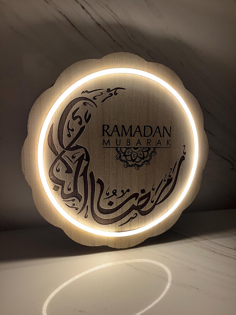 Op de afbeelding: Ronde houten Ramadan Mubarak-bord met een geschulpte rand. Het bevat Arabische kalligrafie en de Engelse woorden "Ramadan Mubarak". Een warm wit licht schijnt rond de rand en benadrukt de tekst.