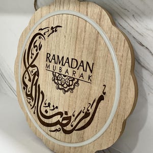 Op de afbeelding: Ronde houten Ramadan Mubarak-bord met een geschulpte rand. Het bord toont de woorden "Ramadan Mubarak" in het Engels en Arabisch kalligrafie, met een decoratief maan- en sterontwerp. Een witte ring omringt het centrale ontwerp.