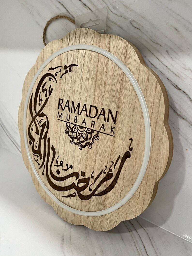 Op de afbeelding: Ronde houten Ramadan Mubarak-bord met een geschulpte rand. Het bord toont de woorden "Ramadan Mubarak" in het Engels en Arabisch kalligrafie, met een decoratief maan- en sterontwerp. Een witte ring omringt het centrale ontwerp.
