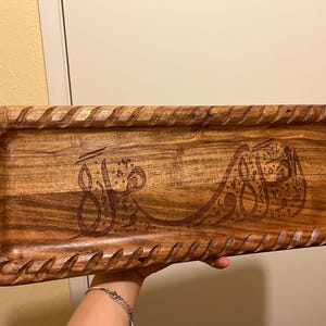 May include: Rectangular wooden serving tray with carved edges and engraved Arabic calligraphy. The tray is a warm brown color and appears to be made of hardwood. The calligraphy is a darker brown, creating a contrast.