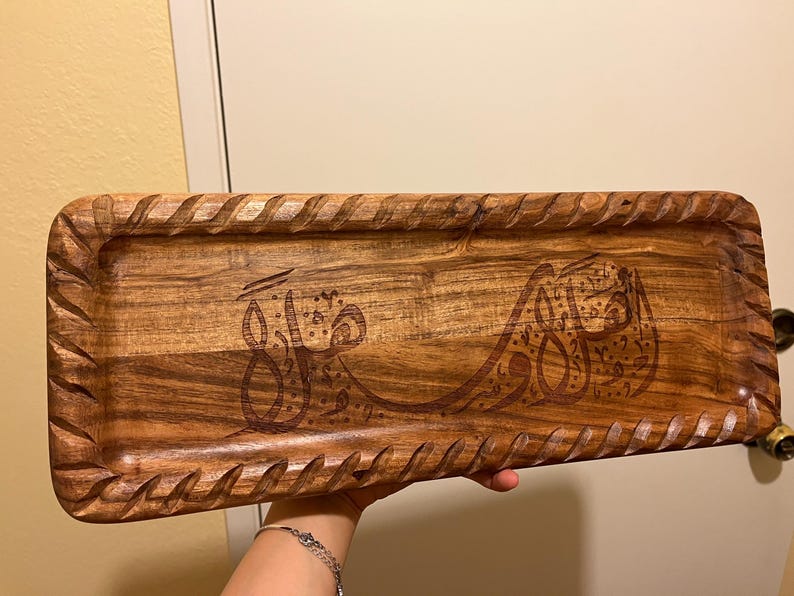 May include: Rectangular wooden serving tray with carved edges and engraved Arabic calligraphy. The tray is a warm brown color and appears to be made of hardwood. The calligraphy is a darker brown, creating a contrast.