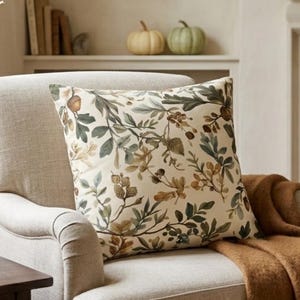 May include: A square throw cushion featuring a cream-coloured background with a botanical print of green and brown oak leaves and acorns. The cushion is positioned on a light-coloured upholstered armchair with a brown throw blanket draped over the side.
