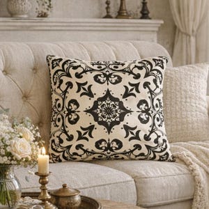 May include: A cozy living room scene featuring a tufted beige sofa adorned with decorative pillows and a cream-colored throw blanket. A round wooden coffee table holds a tray with a vase of white roses, candles, and reading glasses. A lamp and flowers sit on a side table.