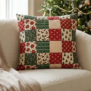 Op de afbeelding: Een vierkant sierkussen met een patchworkontwerp van afwisselende rode, groene en crèmekleurige vierkanten. De stof toont diverse kerstthema-patronen, waaronder kerststerren, zuurstokken, hulstbladeren, sneeuwvlokken en dennenbomen.