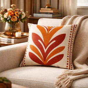 Può includere: Cuscino decorativo quadrato con sfondo crema e un grande motivo floreale stilizzato nei toni dell'arancione e del rosso. Il cuscino ha un bordo a pois in una tonalità arancione più scura ed è posto su una poltrona color crema.