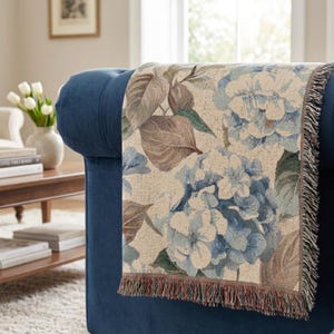 Könnte beinhalten: Eine gewebte Wohndecke mit einem detaillierten Blumenmuster aus blauen Hortensienblüten und großen braunen Blättern auf cremefarbenem Grund. Die rechteckige Decke hat eine dekorative Fransenborte an der Unterkante und liegt über einem dunkelblauen Sessel.