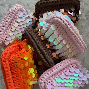 May include: A collection of small, rectangular crochet pouches featuring thick yarn construction and overlapping iridescent sequins. The pouches are available in bright orange, soft pastel pink, and dark brown, each with a distinct, textured knit pattern.