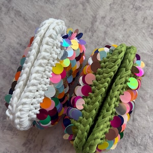 May include: Two crocheted pouches, one in white and one in olive green, both featuring a dense braided texture and covered in numerous round, iridescent, multi-colored sequins that reflect light in shades of pink, orange, blue, and yellow.