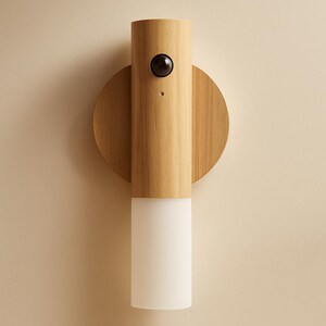May include: A modern wall sconce with a cylindrical wooden body and a circular wooden backplate. The top section features a black sensor, and the bottom has a white, frosted glass light. The sconce is mounted on a beige wall.