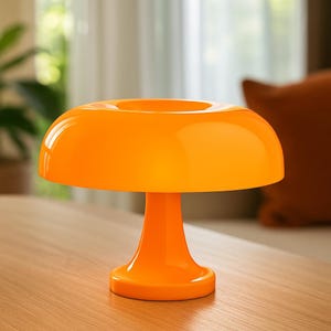 Peut inclure: Une lampe de table orange vif en forme de champignon. La lampe a un abat-jour arrondi avec une ouverture centrale et un pied en forme de piédestal. La finition brillante reflète la lumière, créant une lueur chaleureuse. Luminaire décoratif.