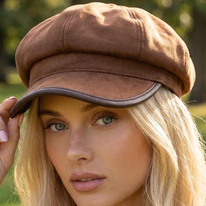 May include: A brown newsboy cap featuring a soft, suede-like texture and a structured crown. The hat includes a curved brim with a dark brown leather-look trim along the edge, providing a distinct contrast to the main body of the headwear.