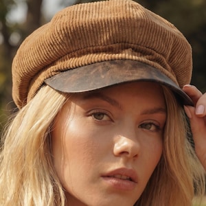 May include: A tan corduroy newsboy cap featuring a textured fabric crown and a smooth, dark brown faux leather brim. The hat has a structured, rounded top with vertical ribbed detailing throughout the fabric, providing a classic, multi-panel design.