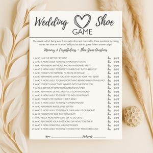 Op de afbeelding: Een crèmekleurige trouwspelkaart met de tekst "Wedding Shoe Game" en een hartafbeelding. De kaart bevat een lijst met vragen over geheugen en vergeetachtigheid, met ruimte om "her" of "him" antwoorden aan te kruisen. Een decoratieve droogbloem en een kleine bal staan op de achtergrond.