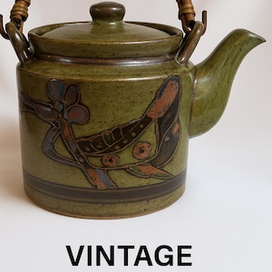 Puede incluir: Tetera japonesa vintage con asa y tapa tejidas. La tetera de cerámica verde oliva presenta un diseño floral en marrón y azul. El texto "VINTAGE JAPANESE TEAPOT" se muestra debajo de la tetera.