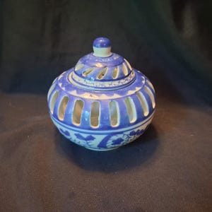 May include: A round ceramic jar with a removable lid, featuring a white base with blue painted floral patterns. The body and lid are designed with vertical cut-out slots around the circumference, and the lid is topped with a small knob.