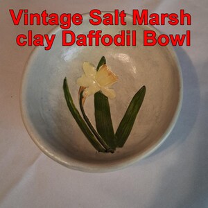 May include: A vintage clay bowl with a daffodil design. The bowl is light gray with a painted daffodil in the center, featuring a yellow flower and green leaves. The text "Vintage Salt Marsh clay Daffodil Bowl" is in red.