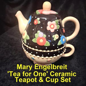 Könnte beinhalten: Keramik-Tee-Set 'Tea for One'. Die schwarze Teekanne ist mit bunten Blumen und weißen Punkten verziert. Das Set enthält eine passende Tasse. Der Text auf dem Bild lautet: Mary Engelbreit 'Tea for One' Ceramic Teapot & Cup Set.