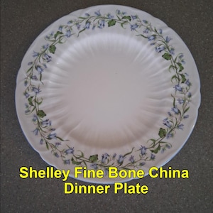 May include: White fine bone china dinner plate with a scalloped edge. The plate is decorated with a floral pattern of blue bellflowers and green leaves around the rim. The text "Shelley Fine Bone China Dinner Plate" is displayed in yellow.