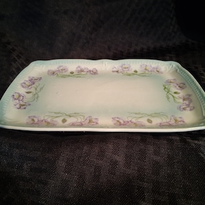 May include: A rectangular porcelain serving tray with a white center and a light blue border. The tray features a delicate floral pattern of purple flowers and green stems arranged along the edges, with a textured, scalloped rim detail.