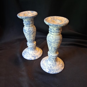 May include: Two ceramic candle holders with a blue and white speckled pattern. The candle holders have a round base, a cylindrical stem, and a flared top. The overall design is rustic and decorative.