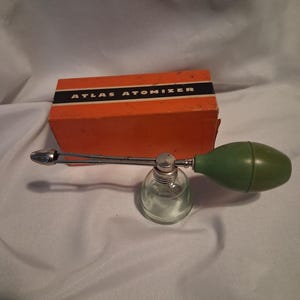 May include: An Atlas Atomizer with a clear glass base, a silver-toned metal nozzle, and a green rubber bulb. The atomizer is positioned in front of its rectangular orange cardboard box, which features the text ATLAS ATOMIZER in black and white.