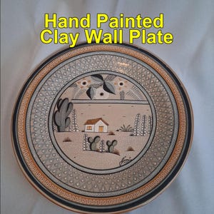 Puede incluir: Plato de pared de arcilla pintado a mano con un diseño de paisaje desértico. El plato presenta una escena central de una casa, cactus y plantas, rodeada de patrones decorativos en tonos beige, marrón y negro.