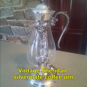 May include: Vintage Sheridan silverplate coffee urn with a clear glass carafe. The silver-colored metal has ornate detailing on the handle, base, and lid. The text "Vintage Sheridan silverplate coffee urn" is displayed in yellow.