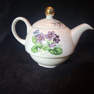 May include: A white ceramic teapot with a lid and handle. The teapot is decorated with purple violets and green leaves. The lid has a gold knob. The teapot is sitting on a black surface.