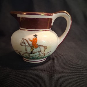 May include: Small, white ceramic pitcher with a brown rim and handle. The pitcher features a hand-painted scene of a person in an orange coat riding a horse. The base and the inside rim are trimmed in a dark red color.