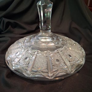 Peut inclure: Un plat de service en verre taillé transparent avec un couvercle. Le couvercle a une poignée facettée en forme de diamant. Le plat présente des motifs complexes en forme d'étoiles et géométriques. Le verre a une qualité brillante et réfléchissante.