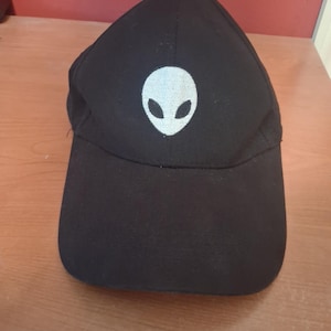 May include: A black baseball cap featuring a white embroidered alien head icon centered on the front panel. The cap has a structured crown, a curved brim, and a button at the top center. The design is monochromatic with a simple, clean aesthetic.