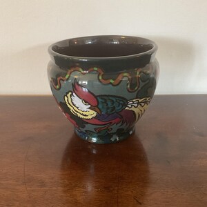 May include: A ceramic planter with a dark brown interior and a teal exterior. The planter features a colourful pheasant design with red, yellow, and blue accents. The rim is decorated with a green and orange pattern.