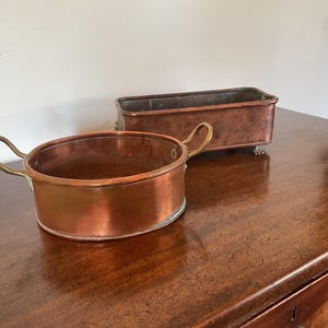 May include: Two copper containers on a wooden surface. One is round with brass handles, the other is rectangular with small feet. Both have a warm, reddish-brown hue, suggesting age and use. The containers are likely for cooking or storage.