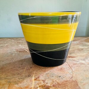 May include: A tapered ceramic planter featuring horizontal bands of bright yellow, olive green, and black. The exterior surface is decorated with thin, intersecting white lines that wrap around the vessel in a continuous, angular pattern.