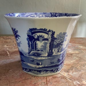 May include: A cream-coloured ceramic planter featuring a blue transferware pattern. The design depicts architectural ruins, trees, and grazing cattle in a pastoral landscape. The interior rim is decorated with a repeating floral and scroll motif.