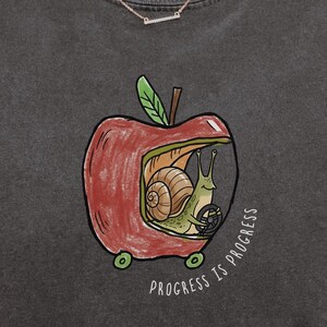May include: Dark gray t-shirt featuring a whimsical graphic of a snail driving a car made from a red apple. The apple has green wheels and a green leaf stem. The text "PROGRESS IS PROGRESS" is printed below the apple.