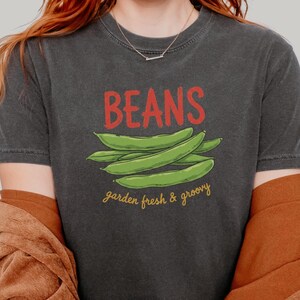 May include: Dark gray t-shirt with the word "BEANS" in red above a graphic of green beans. The text "garden fresh & groovy" is below the beans. The person is wearing a brown corduroy jacket and blue jeans.