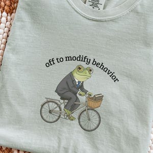 May include: A light green t-shirt with the text "off to modify behavior." The shirt features a cartoon frog in a suit riding a bicycle with a basket. The frog is holding a book.