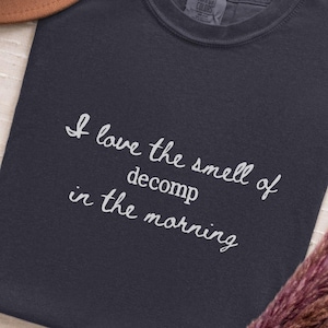 May include: Graphite-colored t-shirt with the text "I love the smell of decomp in the morning" in white script. The shirt is displayed with a brown hat and dried floral arrangement.