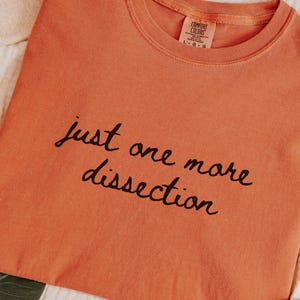 May include: An orange t-shirt with the phrase "just one more dissection" in black script. The shirt is folded and lies on a cream-colored surface, with a hat and other accessories visible.