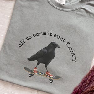 May include: Grey t-shirt with a black crow graphic on a skateboard, wearing red sneakers. The text "off to commit aunt foolery" is arched above the crow. The word "GREY" is in the upper left corner.