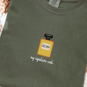 May include: A moss green t-shirt with a graphic of a yellow perfume bottle that says "DECOMP" with the words "my signature scent" below it. The shirt is folded and lies on a woven rug.
