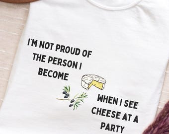 May include: White t-shirt with the text "I'M NOT PROUD OF THE PERSON I BECOME WHEN I SEE CHEESE AT A PARTY." The shirt features a cheese illustration and olive branch graphic. A brown hat is visible in the background.