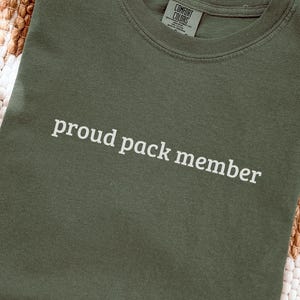 May include: A moss green t-shirt with the words "proud pack member" printed in white. The shirt is made of a soft material and has a crew neck. The Comfort Colors tag is visible at the neckline.