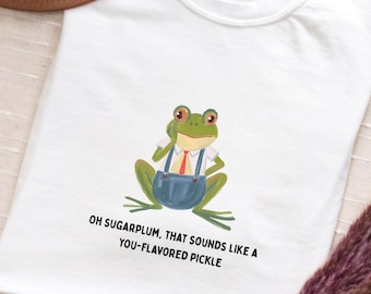 May include: White t-shirt featuring a cartoon frog wearing a shirt, tie, and overalls. The frog is green with red eyes. Text on the shirt reads, "OH SUGARPLUM, THAT SOUNDS LIKE A YOU-FLAVORED PICKLE."