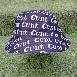 Puede incluir: Sombrero de pescador negro con la palabra "Cunt" en blanco repetida. El sombrero se muestra sobre un soporte de metal negro, sobre un fondo de césped artificial verde.